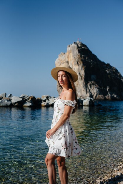 Una hermosa mujer joven con un sombrero y un vestido ligero está caminando por la orilla del océano con el telón de fondo de enormes rocas en un día soleado. Viajes de turismo y vacaciones.