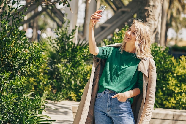 Hermosa mujer joven sonriendo mientras sostiene el teléfono inteligente paseando al aire libre en la ciudad Mujer feliz tomando selfie en el teléfono móvil Chica bonita haciendo autorretrato con cara divertida en el teléfono celular Viajes