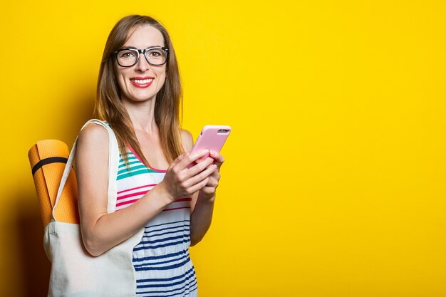 Hermosa mujer joven sonriente con karimat en una bolsa sosteniendo un teléfono sobre un fondo amarillo.