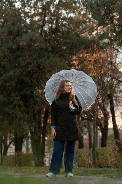 Hermosa mujer joven sonriente bajo un paraguas transparente en el parque de otoño Chica en abrigo negro camina sobre la lluvia