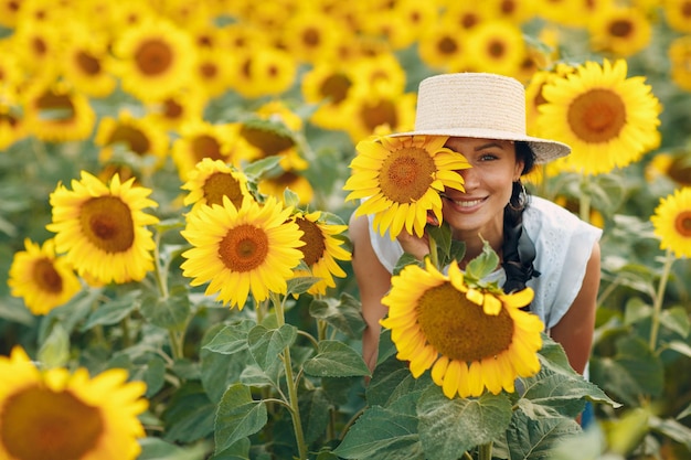 Hermosa mujer joven sonriente con un sombrero con flor en los ojos y la cara en un campo de girasoles