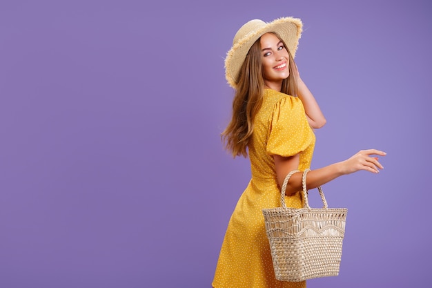 Hermosa mujer joven sonriente en vestido amarillo bolso de sombrero de paja de verano aislado sobre fondo violeta pastel