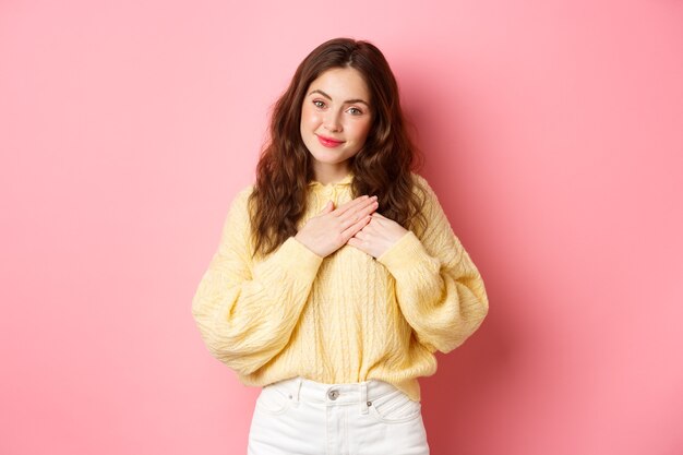 Hermosa mujer joven con una sonrisa amable, tomados de la mano en el corazón y sonriendo, recibir cumplidos, sentirse sincero, estar agradecido, decir gracias, de pie sobre una pared rosa.