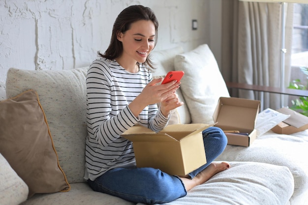 Foto hermosa mujer joven está sosteniendo una caja de cartón y desempacando un teléfono inteligente sentado en el sofá en casa