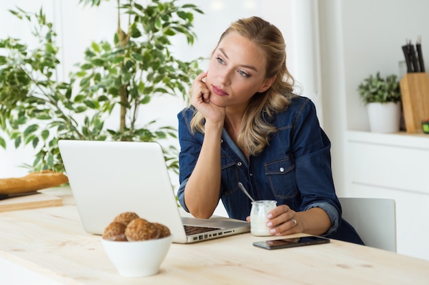 Hermosa mujer joven con su computadora portátil y comer yogur en casa.