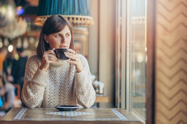 Hermosa mujer joven en suéter caliente en el café cerca de la ventana con café. Acogedora mañana de otoño o invierno.