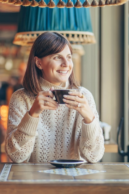 Hermosa mujer joven en suéter caliente en el café cerca de la ventana con café. Acogedora mañana de otoño o invierno.