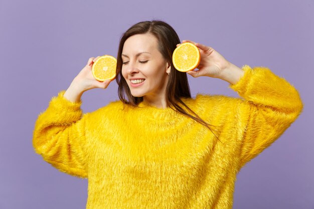 Hermosa mujer joven en suéter de piel manteniendo los ojos cerrados sostiene mitades de fruta fresca de naranja madura aislada sobre fondo violeta pastel. Estilo de vida vivo de la gente, concepto de vacaciones relajantes. Simulacros de espacio de copia.