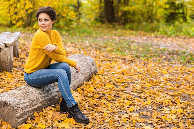 Hermosa mujer joven en un suéter de punto amarillo y jeans azules se sienta en un tronco en un parque de otoño