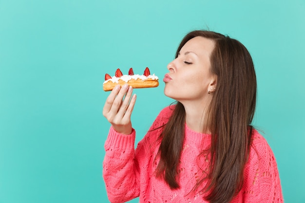 Foto hermosa mujer joven en suéter rosa de punto con los ojos cerrados sosteniendo en la mano comiendo soplando enviando aire beso a pastel de eclair aislado sobre fondo azul. concepto de estilo de vida de personas. simulacros de espacio de copia.