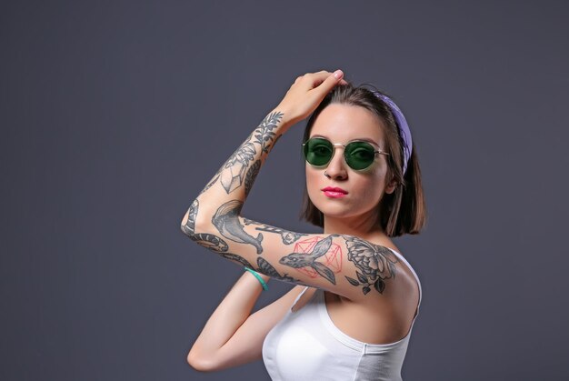 Foto hermosa mujer joven con tatuaje con gafas de sol y posando sobre fondo gris