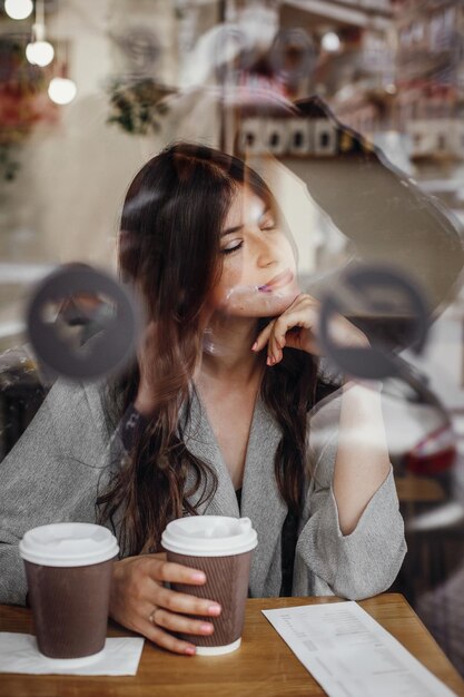 Foto hermosa mujer joven con una taza de café sentada en la cafetería y relajándose en la ventana en la calle de la ciudad elegante chica hipster con un cabello hermoso disfrutando del tiempo con café