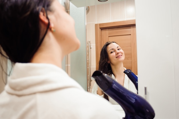 Foto hermosa mujer joven en toalla de baño está utilizando un secador de pelo y sonriendo mientras se mira en el espejo en el baño.