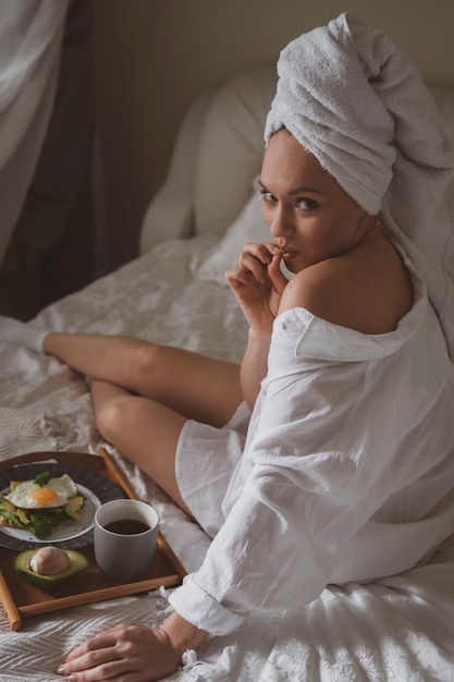 Hermosa mujer joven con una toalla en la cabeza en la cama come un desayuno saludable y se lame los dedos