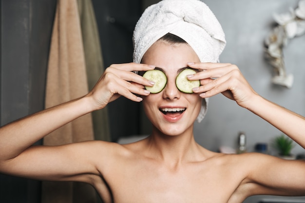 Hermosa mujer joven con una toalla envuelta alrededor de su cabeza sosteniendo rodajas de pepino en su cara en el baño