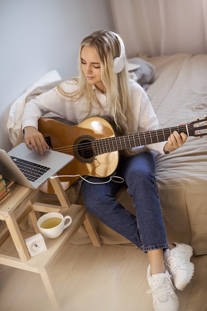 Hermosa mujer joven tocando la guitarra mientras está sentado en la cama en su casa. Retrato de linda chica en