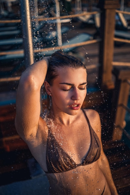 Hermosa mujer joven en traje de baño con una figura perfecta en la ducha a la luz del atardecer junto al mar Chica con bronceado dorado de vacaciones