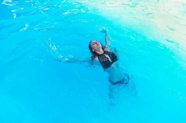 Hermosa mujer joven en traje de baño negro nadando en la piscina azul sobre su espalda Joven nadadora en el complejo vacacional Actividad deportiva concepto de salud sonrisa en la cara