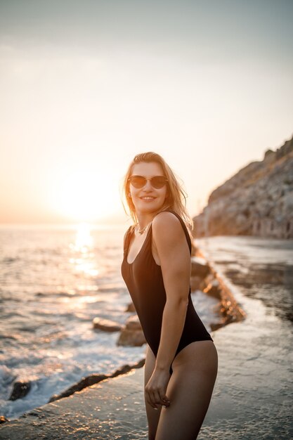 Hermosa mujer joven en traje de baño negro sola en la playa junto al mar al atardecer. Una esbelta niña adulta descansando en la orilla del mar al atardecer. Concepto de vacaciones de verano. Enfoque selectivo