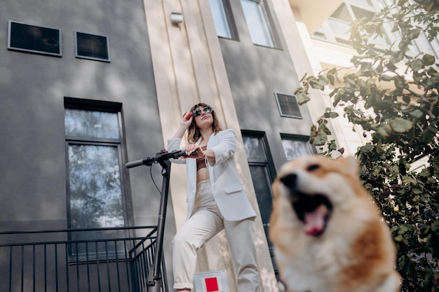 Hermosa mujer joven en traje blanco de pie con su scooter eléctrico y perro COrgi cerca de un edificio moderno en la ciudad y mirando hacia otro lado