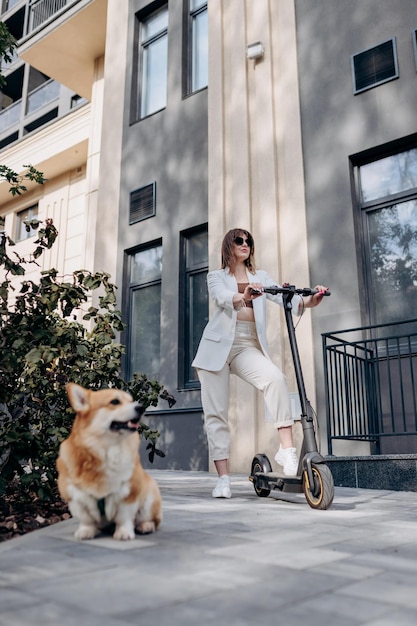 Hermosa mujer joven en traje blanco de pie con su scooter eléctrico y perro COrgi cerca de un edificio moderno en la ciudad y mirando hacia otro lado