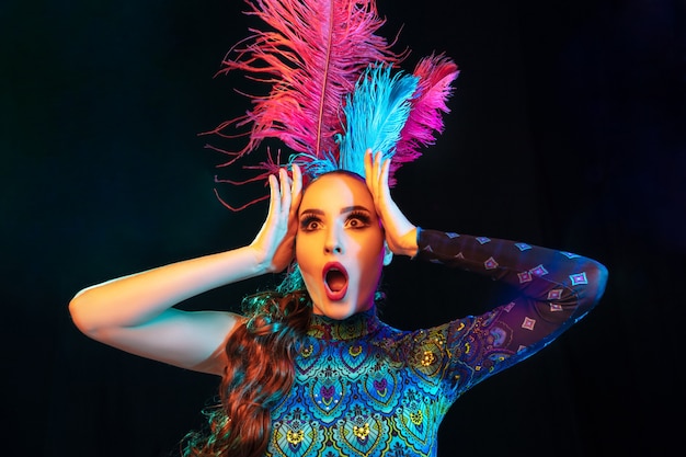 Hermosa mujer joven en traje de carnaval y mascarada en coloridas luces de neón en pared negra