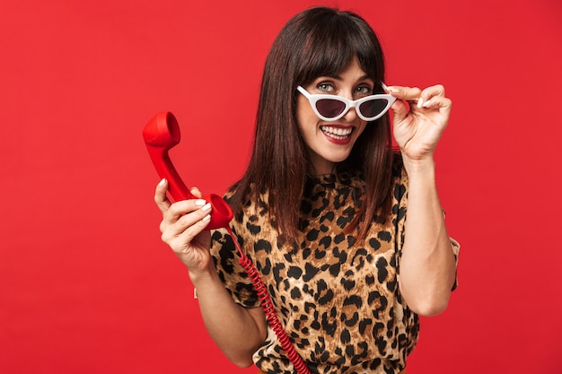 Hermosa mujer joven vestida con camiseta estampada de animales posando aislada sobre pared roja con gafas de sol hablando por teléfono.