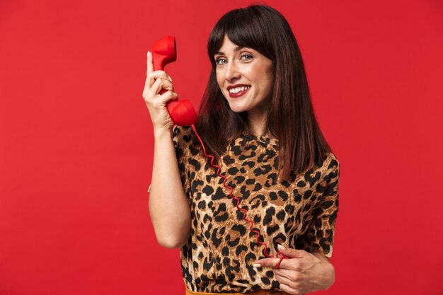 Hermosa mujer joven vestida con camiseta estampada de animales posando aislada sobre pared roja hablando por teléfono.