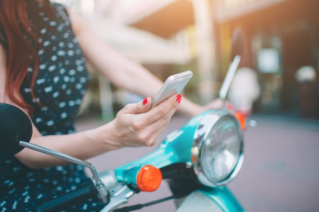 Hermosa mujer joven vestida casual sentado en scooter y teléfono Holdind. Niña sentada en la bicicleta y utilizando teléfono inteligente celular