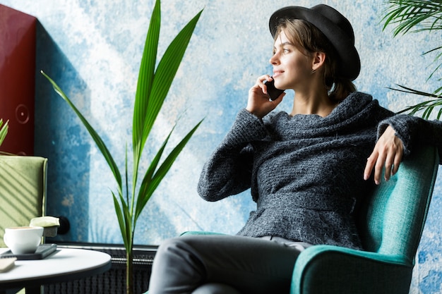 Hermosa mujer joven vestida con suéter y sombrero sentado en una silla en la mesa de café, hablando por teléfono móvil, interior elegante