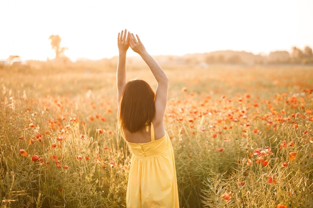 Hermosa mujer joven con un vestido amarillo caminando en un campo de amapolas en un día de verano. Chica disfrutando de flores en el campo. Enfoque selectivo