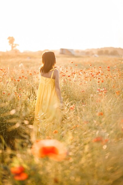 Hermosa mujer joven con un vestido amarillo caminando en un campo de amapolas en un día de verano. Chica disfrutando de flores en el campo. Enfoque selectivo