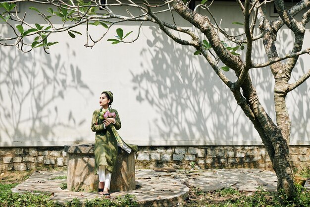 Hermosa mujer joven en vestido de ao dai sentado en el jardín con ramo de flores de loto y mirando a otro lado