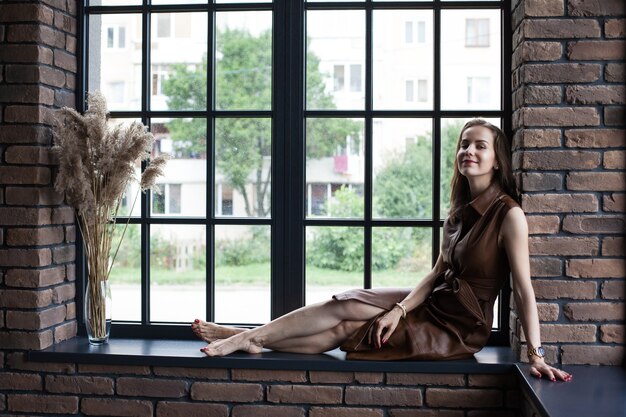 Foto hermosa mujer joven en vestido de cuero marrón se sienta en el alféizar de la ventana descalzo