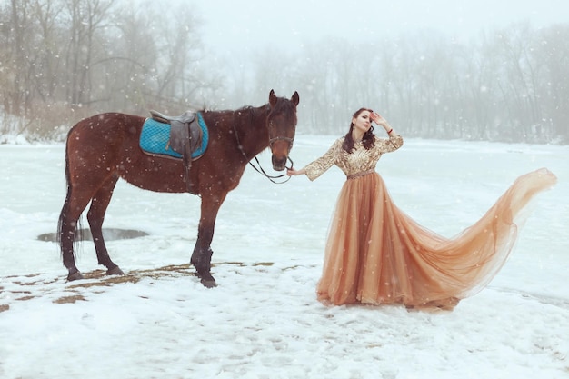 Hermosa mujer joven en un vestido elegante de pie junto a un lago congelado Ella sostiene un caballo para las riendas