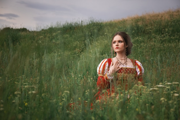 Hermosa mujer joven con un vestido medieval rojo largo está sentada en la hierba en el campo