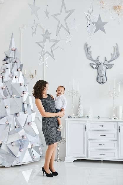 Hermosa mujer joven en un vestido de moda posando con su pequeño y lindo hijo en el interior decorado para Navidad