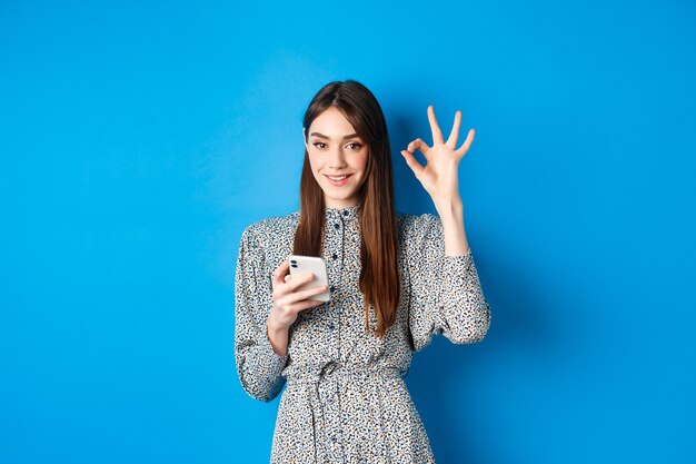 Hermosa mujer joven en vestido mostrando signo bien y con smartphone, sonriendo a la cámara, de pie sobre azul.