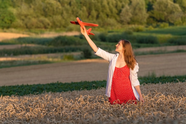 Hermosa mujer joven en vestido rojo y camisa blanca con avión de juguete en la mano Concepto de viajes aéreos Campo de trigo