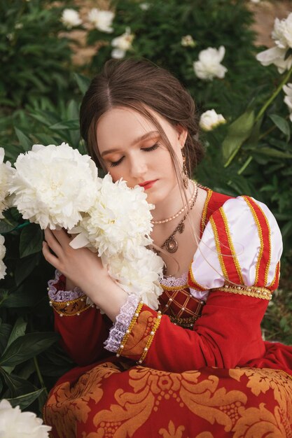 Hermosa mujer joven con un vestido rojo medieval sostiene peonías blancas