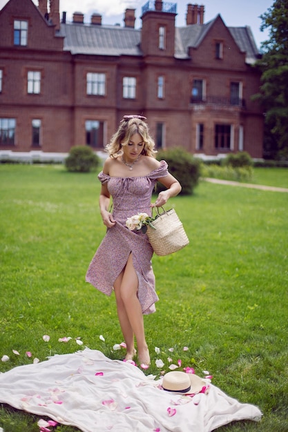 Hermosa mujer joven con un vestido rosa se encuentra en un prado con una canasta con flores rosas en el fondo de un castillo en verano