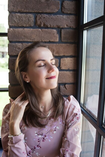 Hermosa mujer joven en vestido rosa se sienta en el alféizar de la ventana