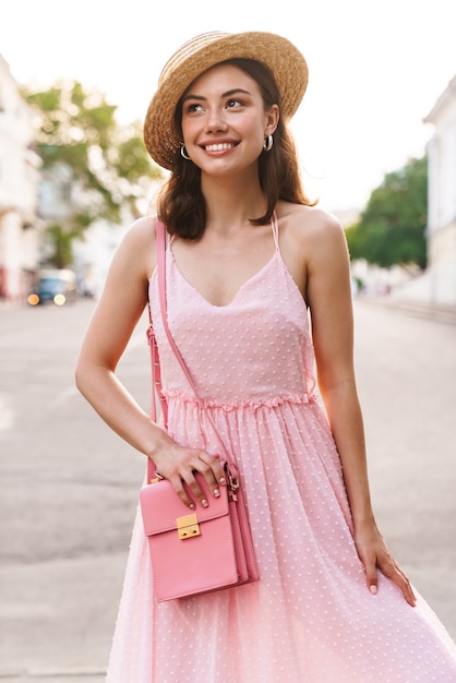 Hermosa mujer joven con vestido de verano y sombrero de paja sonriendo mientras camina por las calles de la ciudad