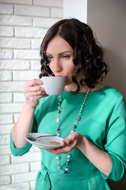 Hermosa mujer joven en vestido verde de pie en una pared de ladrillos con una taza de café blanco en la mano