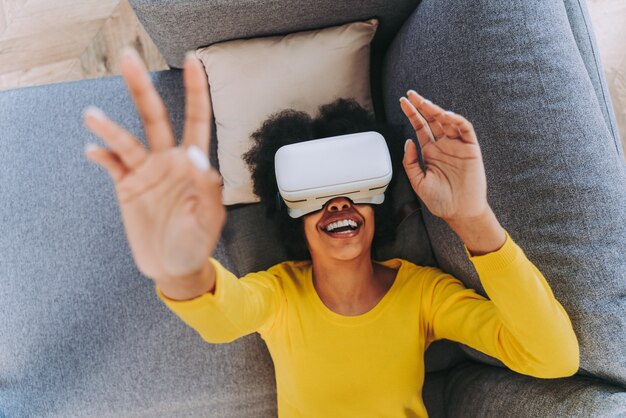 Hermosa mujer jugando en casa con auriculares VR - Mujer negra acostada en el sofá mientras interactúa con tecnología de realidad aumentada