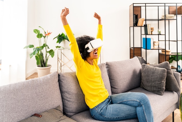 Hermosa mujer jugando en casa con auriculares VR - Mujer negra acostada en el sofá mientras interactúa con tecnología de realidad aumentada