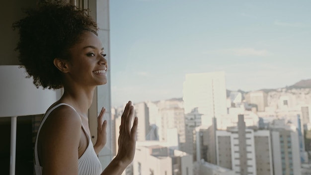 Hermosa mujer latina despertando por la mañana, el sol brilla sobre ella desde la ventana grande Una joven feliz saluda el nuevo día con la cálida luz del sol y el paisaje de la ciudad en la ventana