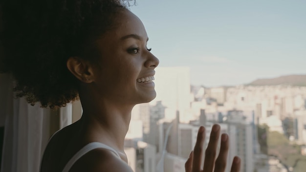 Hermosa mujer latina despertando por la mañana el sol brilla sobre ella desde la ventana grande Mujer joven feliz saluda el nuevo día con la cálida luz del sol y el paisaje de la ciudad en la ventana