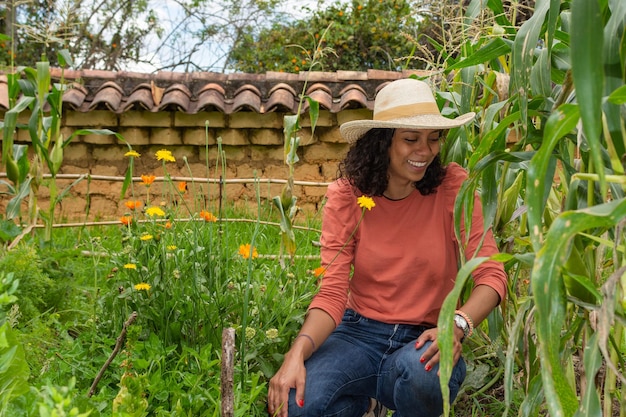 Hermosa mujer latina con sombrero y jean azul en el jardín de su casa rodeada de flores medicinales y plantas de maíz mientras sonríe