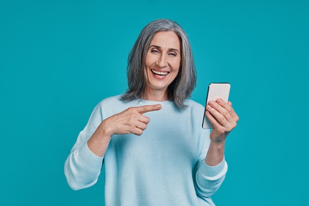 Hermosa mujer madura apuntando teléfono inteligente y sonriendo mientras está de pie contra el fondo azul.